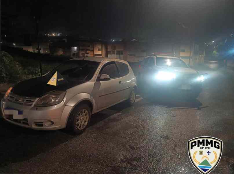 Imagem de um carro branco estacionado à noite, próximo a um veículo da polícia com faróis acesos, em uma área urbana de Nova Serrana.