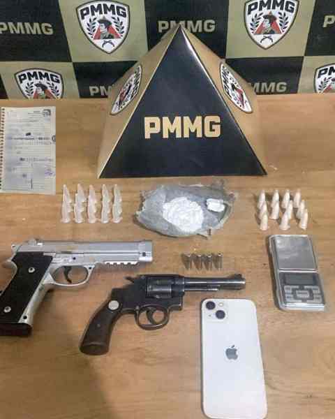 Uma mesa com um bloco de anotações, uma balança de precisão, um revólver, uma pistola, pinos vazios de plástico, uma barra de substância semelhante à cocaína e um celular, em frente a uma parede com o logotipo da Polícia Militar de Minas Gerais.