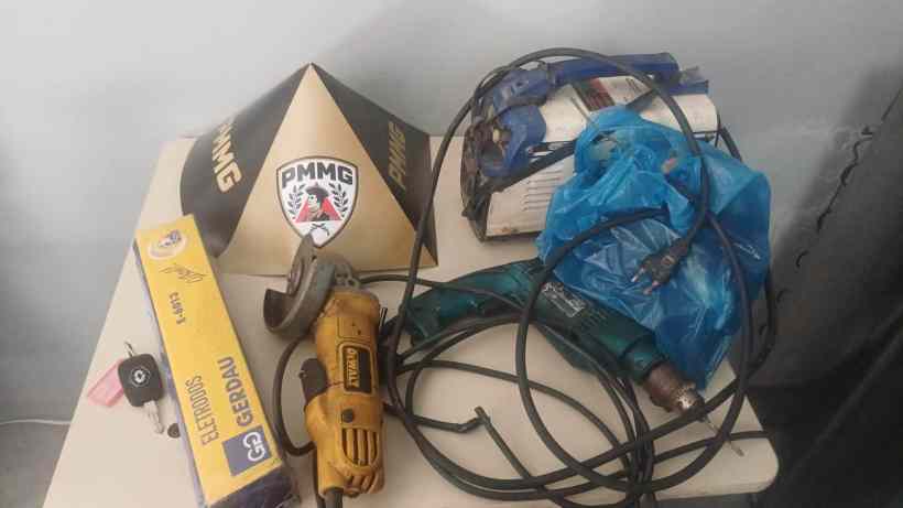 Mesa com ferramentas e objetos recuperados, incluindo uma serra, um veículo de solda, uma caixa azul, e um cabo elétrico, sob um boné da Polícia Militar de Minas Gerais.