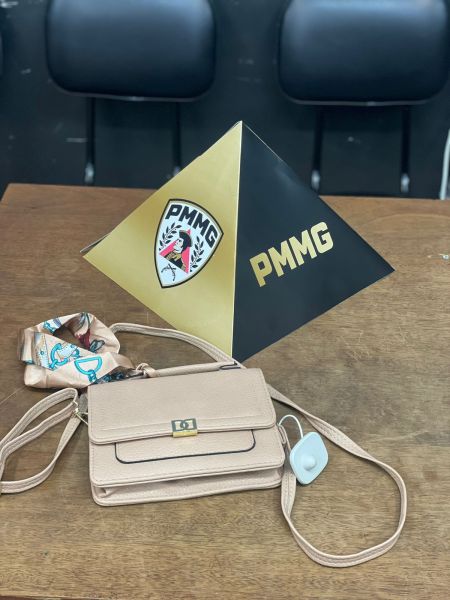 Uma bolsa feminina com etiqueta antifurto e um objeto em formato de pirâmide representando a Polícia Militar de Minas Gerais (PMMG) sobre uma mesa de madeira.