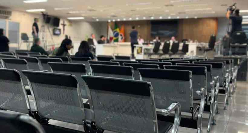 Visão do plenário da Câmara Municipal, com cadeiras vazias e algumas pessoas presentes, decorado com bandeiras do Brasil e do município.