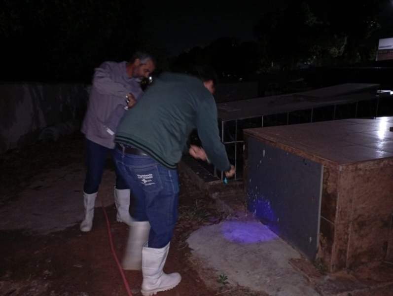 Dois homens usando botas brancas trabalham à noite, um deles usa uma lanterna e o outro aplica uma solução em uma superfície ao redor de um objeto em um ambiente externo.