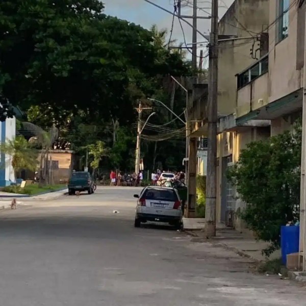 Rua tranquila com árvores e edifícios ao fundo, veículos estacionados e um grupo de pessoas ao longe.