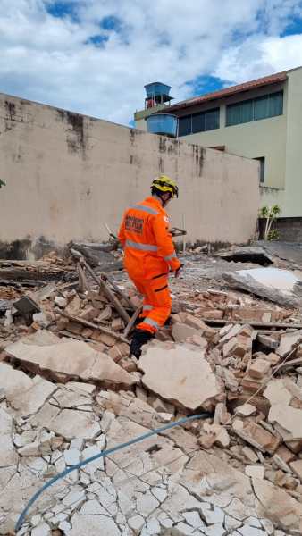Um trabalhador de resgate, vestido com um uniforme laranja e capacete, caminha sobre escombros de uma construção danificada, com um céu parcialmente nublado ao fundo.