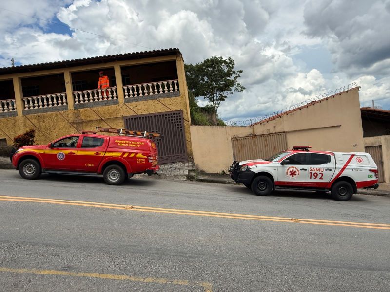 Veículo de bombeiros e ambulância do SAMU estacionados na rua, com um homem em uniforme laranja observando do andar superior de um prédio.