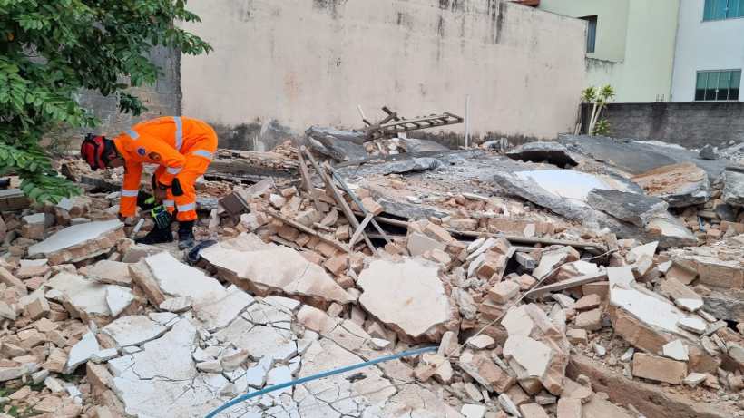 Um trabalhador de resgate, vestido com um uniforme laranja, inspeciona os escombros de um edifício desmoronado, cercado por uma parede e árvores ao fundo.