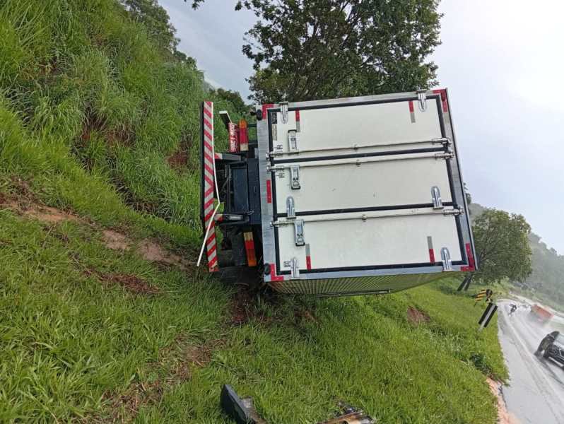 Caminhão tombado na estrada, com o fundo inclinado em uma área de grama e árvores ao redor.