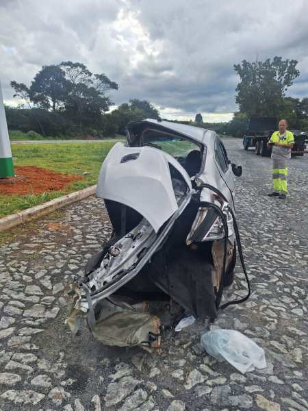 Carro capotado com a parte traseira danificada em uma estrada, com um homem ao fundo observando o acidente.
