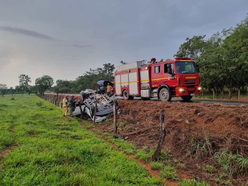Caminhão de bombeiros próximo a um carro capotado em uma estrada, com equipes de resgate trabalhando na cena.