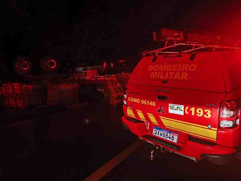 Veículo do Corpo de Bombeiros Militar em operação, com sirenes acionadas, em uma estrada à noite, ao lado de um caminhão tombado e cargas espalhadas.