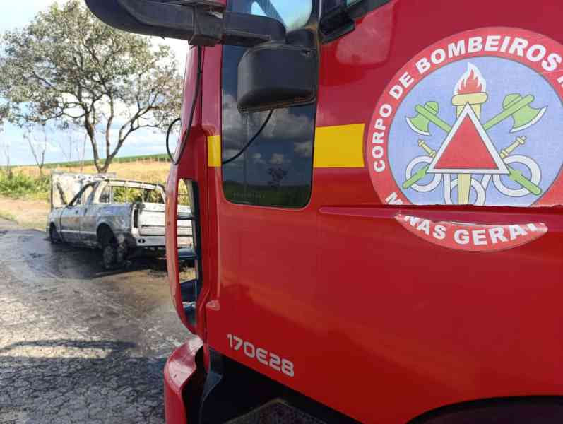 Caminhão do Corpo de Bombeiros com logotipo, em primeiro plano, e veículo queimado ao fundo, em um cenário rural.