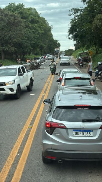Acidente de trânsito em uma estrada, com veículos parados e pessoas na cena, em meio a árvores ao fundo.