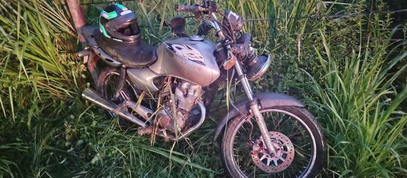 Moto esportiva cinza abandonada em meio a grama alta, com um capacete verde ao lado.