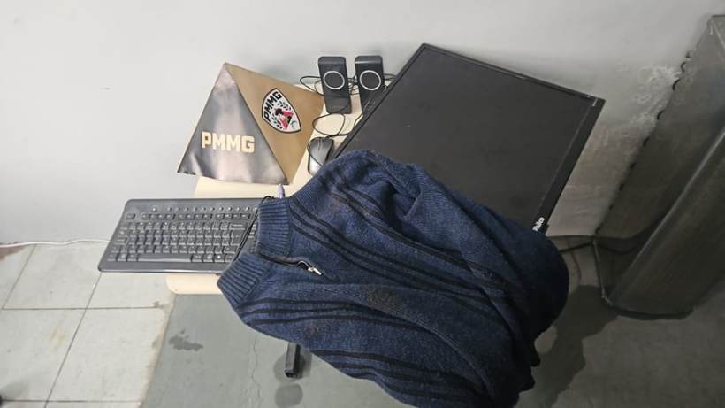 Mesa com um computador, teclado, monitor, alto-falantes e um agasalho encostado, ao fundo uma pasta com o logo da PMMG.