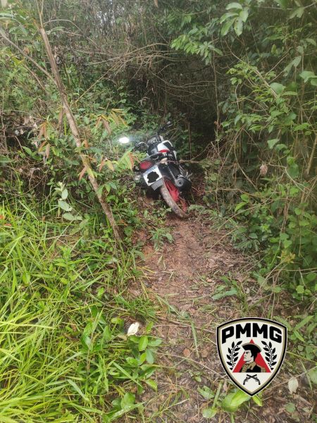 Moto preta e vermelha abandonada em uma trilha cercada por vegetação densa.