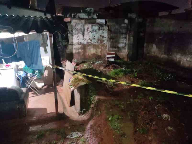 Vista noturna de um espaço exterior com uma área verde, cercada por muros desgastados e um local de descanso com um sofá. Há uma cadeira e uma fita amarela de sinalização no ambiente.