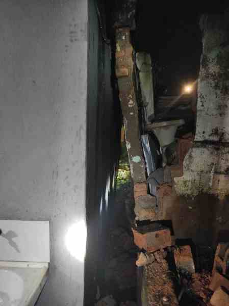 Visualização noturna de uma parede parcialmente derrubada, com tijolos expostos ao lado de uma estrutura de edifício, iluminada por uma luz frontal.