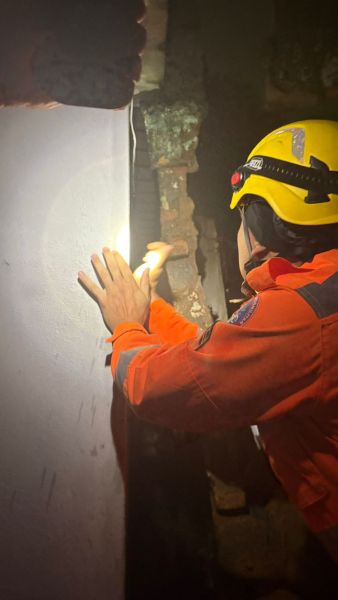 Pessoa usando capacete e traje de resgate, examinando uma parede com uma lanterna em ambiente escuro.