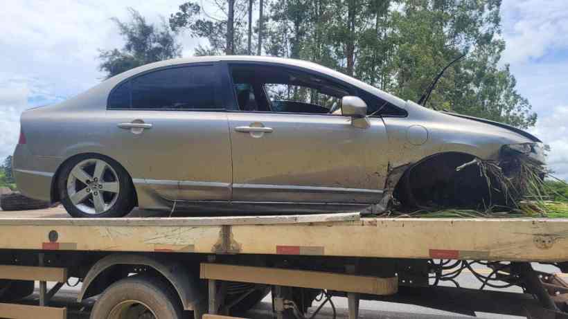 Um carro prata danificado sendo transportado em uma plataforma, com vegetação ao redor.