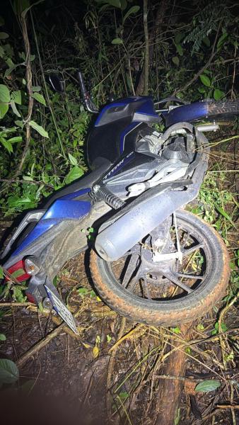 Uma motocicleta caída em uma área de vegetação densa à noite, com detalhes visíveis de barro na roda traseira.