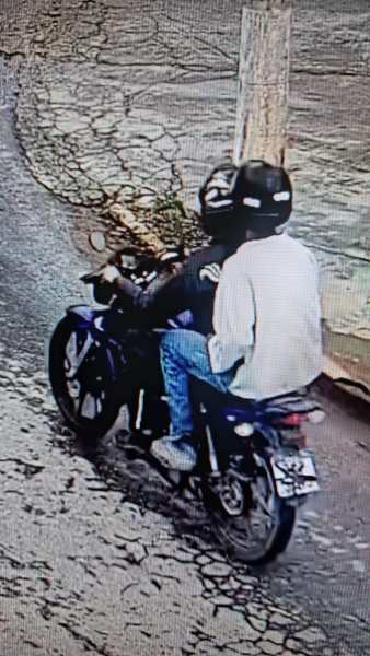 Duas pessoas em uma motocicleta, uma delas usando capacete preto e a outra com uma camiseta branca, enquanto se afastam em uma rua pavimentada.