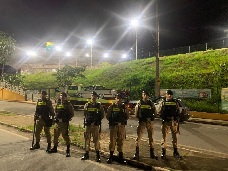 Policiais em pé em uma rua durante a noite, com iluminação forte ao fundo e uma bandeira do Brasil visível.