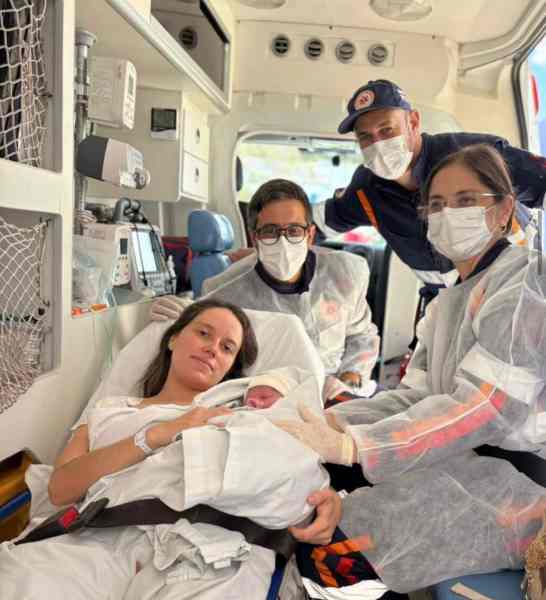 Mulher segurando um bebê recém-nascido em uma ambulância, cercada por profissionais de saúde, todos usando máscaras e equipamentos de proteção.