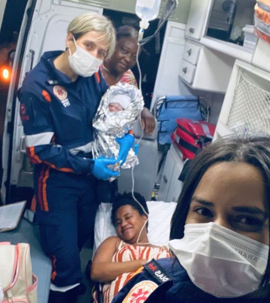 Equipes de paramédicos em uma ambulância, com uma mãe segurando seu recém-nascido envolto em um cobertor prateado, enquanto outra mulher sorri e relaxa ao lado.