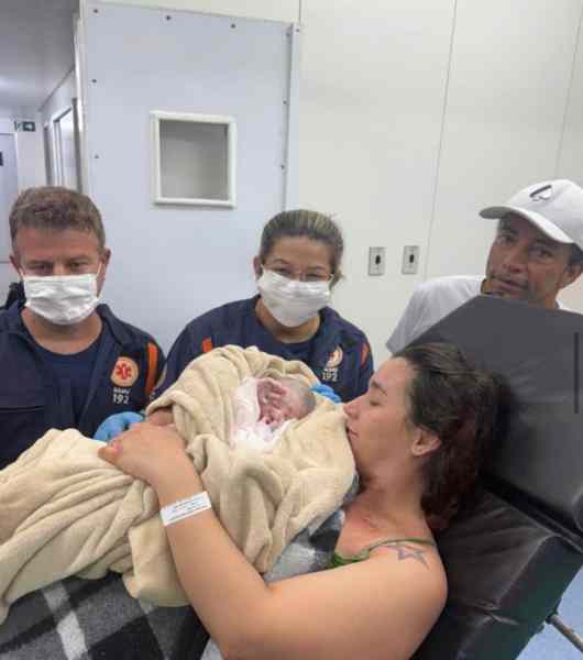 Uma mãe segurando seu bebê recém-nascido envolto em uma manta, cercada por profissionais de saúde e um homem ao fundo, todos usando máscaras em um ambiente hospitalar.