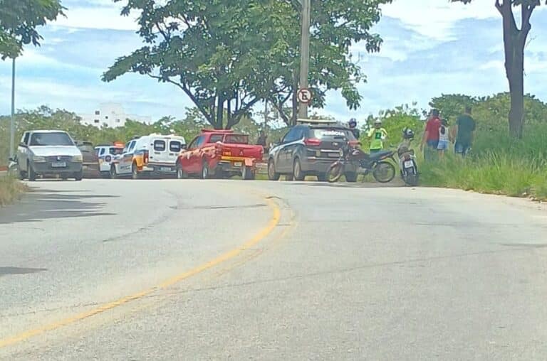 Caminho curvo com veículos estacionados, incluindo um carro vermelho, uma motocicleta e carros de polícia. Há pessoas próximo ao local.