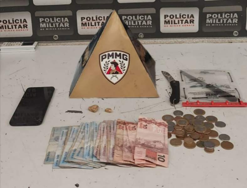 Imagem de uma mesa com itens apreendidos pela polícia, incluindo uma pirâmide com o logo da Polícia Militar de Minas Gerais, um celular preto, uma faca, notas de dinheiro e moedas.