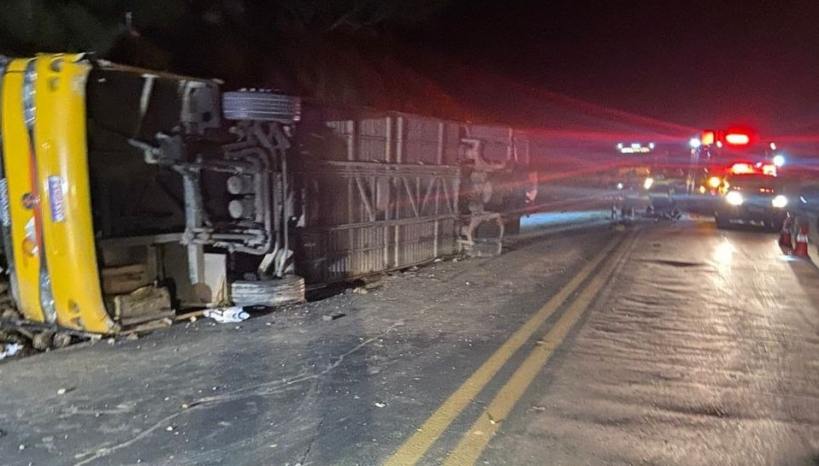 Caminhão acidentado de lado na estrada à noite, com viaturas de emergência ao fundo.