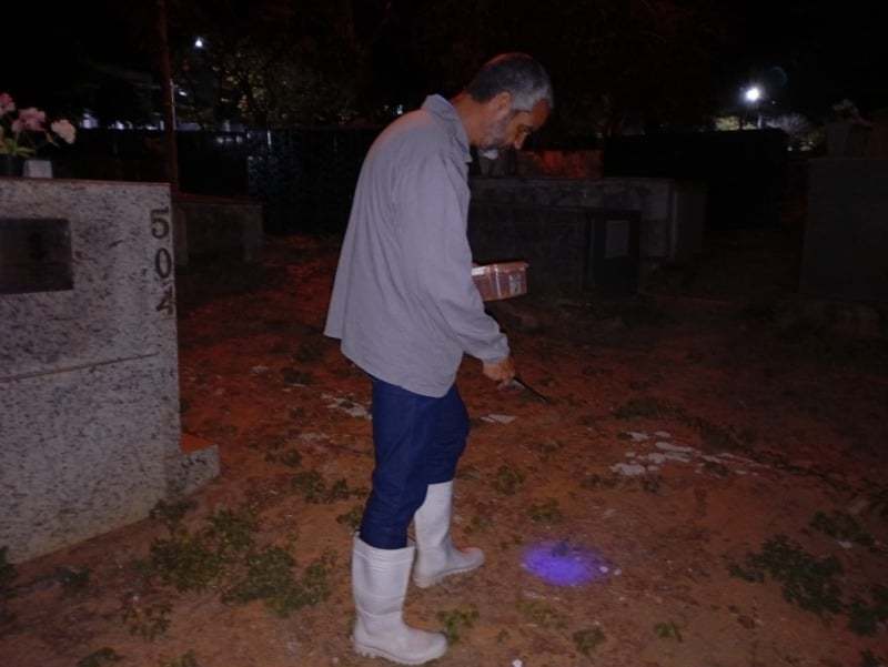 Homem em um cemitério à noite, usando botas brancas e segurando uma caixa enquanto observa o chão iluminado por uma luz roxa.