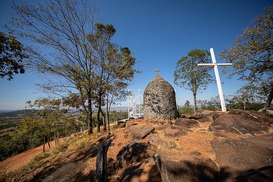 Vista de uma colina com uma estrutura de pedra em forma de cúpula e uma cruz branca ao lado, rodeada por árvores e rochas.