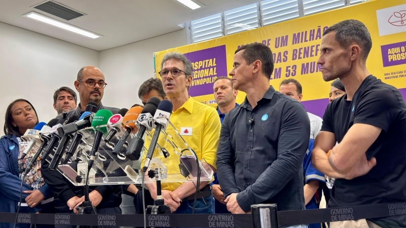 Coletiva de imprensa com várias pessoas, incluindo um homem de camisa amarela falando ao microfone, em frente a um painel sobre o hospital regional de Varginópolis.