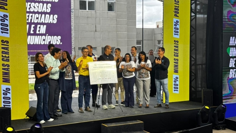 Evento com várias pessoas em um palco, celebrando o lançamento de um projeto, com um painel ao fundo refletindo a mensagem sobre 'Minas Gerais 100% Público'.