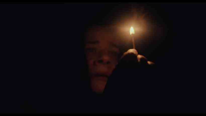 Pessoa segurando uma vela acesa em um ambiente escuro, com uma expressão de medo ou preocupação.
