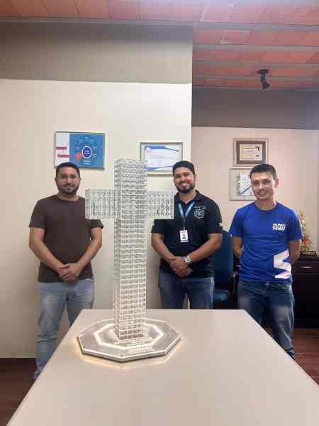 Três homens posando ao lado de uma escultura em forma de cruz em uma mesa de escritório, com certificações e documentos na parede ao fundo.