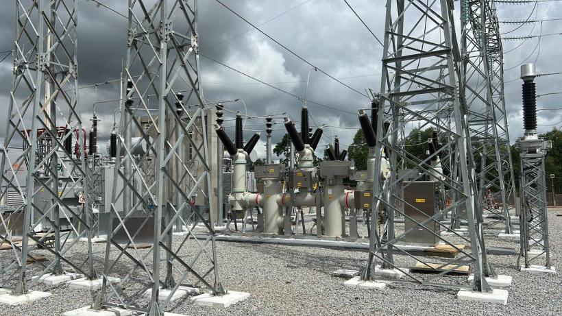 Estação de energia elétrica com torres e equipamentos de conexão, incluindo transformadores e isoladores, sob um céu nublado.
