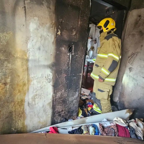 Bombeiro em um ambiente danificado por incêndio, diante de uma parede que apresenta sinais de queima e bagunça no chão com roupas espalhadas.