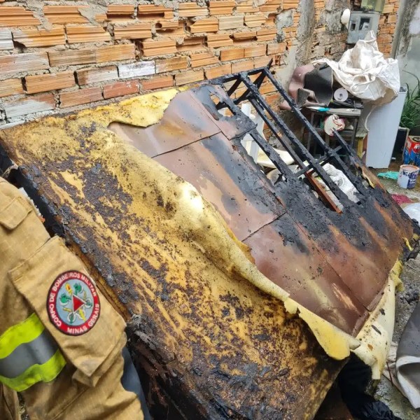 Superfície queimada de um objeto retangular, com metal derretido e camada de espuma amarela, em um ambiente bagunçado com tijolos ao fundo.