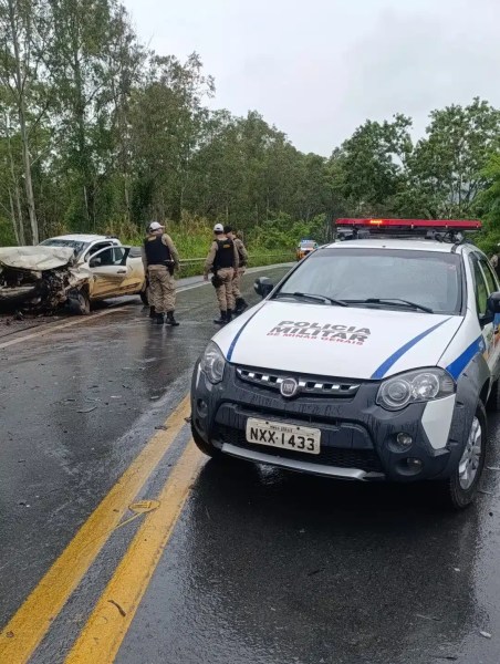 Veículo da polícia em uma estrada, com agentes analisando um carro acidentado ao fundo em uma área arborizada.
