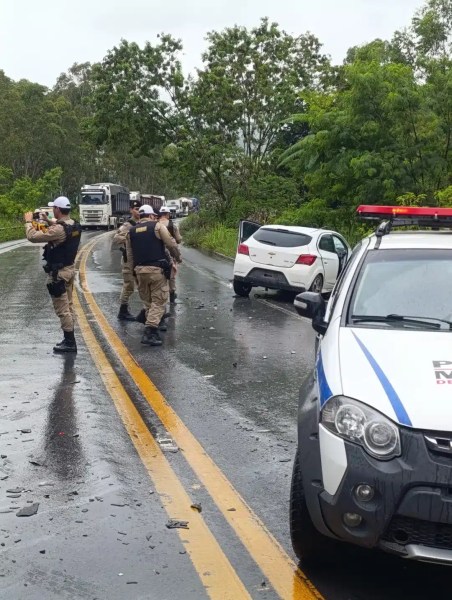 Grupo de policiais em uma estrada, com um carro branco parado e um veículo policial próximo. A pista está molhada e há caminhões ao fundo.