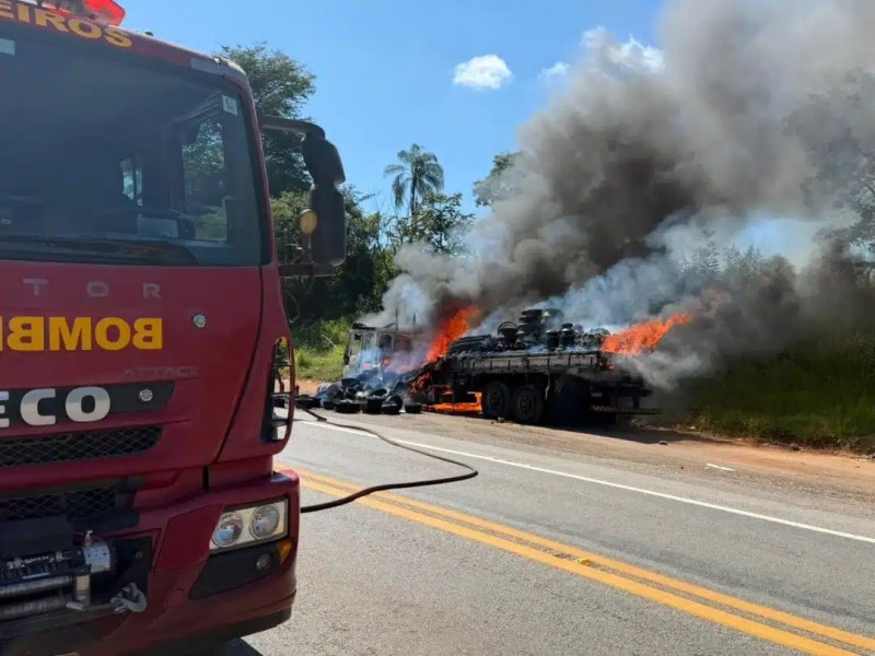 Caminhão dos bombeiros em primeiro plano, com fogo e fumaça saindo de um caminhão em chamas na estrada.
