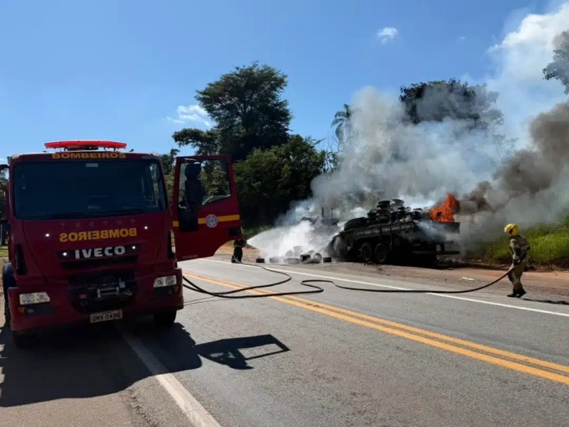 Caminhão dos bombeiros ao lado de um veículo em chamas, com fumaça saindo e bombeiros combatendo o incêndio em uma estrada.