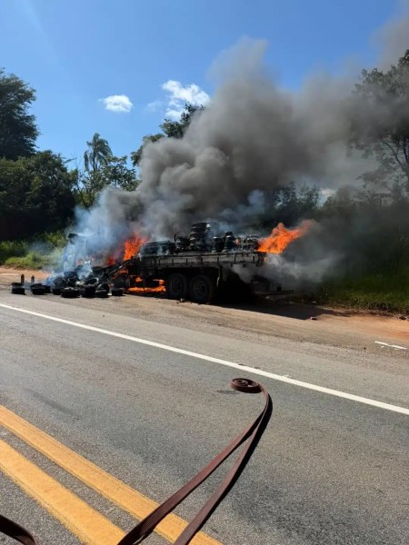 Caminhão em chamas na beira da estrada, com fumaça negra saindo e pneus queimando.