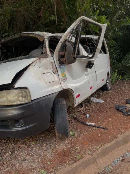 Van branca danificada estacionada ao lado da estrada, com a porta dianteira aberta e danos visíveis na carroceria.