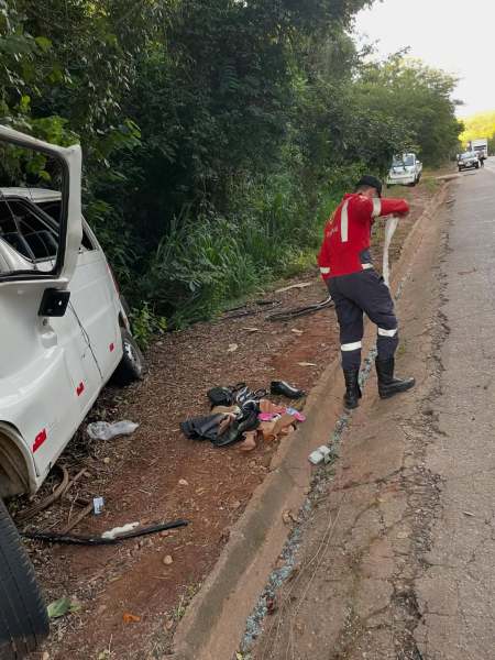 Um homem vestido com uniforme vermelho e preto está em pé ao lado de uma van danificada, enquanto examina a área ao longo da estrada. Roupas e objetos pessoais estão jogados no chão próximo a ele.