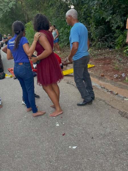 Grupo de pessoas em uma rua, incluindo mulheres em vestidos, um homem com cabelo grisalho e uma pessoa caída ao fundo. O ambiente é ao ar livre, com vegetação ao redor.