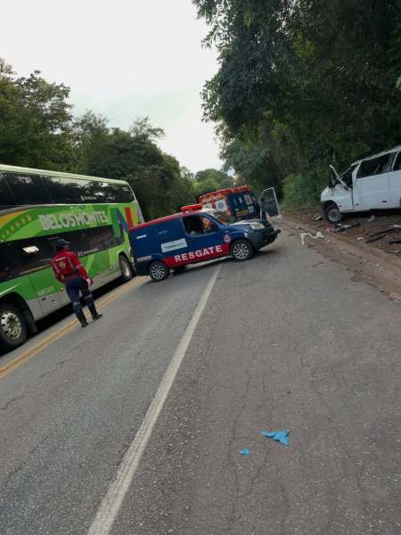 Acidente de trânsito em uma estrada, com um ônibus verde estacionado ao lado de um veículo de resgate e uma van branca danificada. Um homem em uniforme de resgate observa a cena.